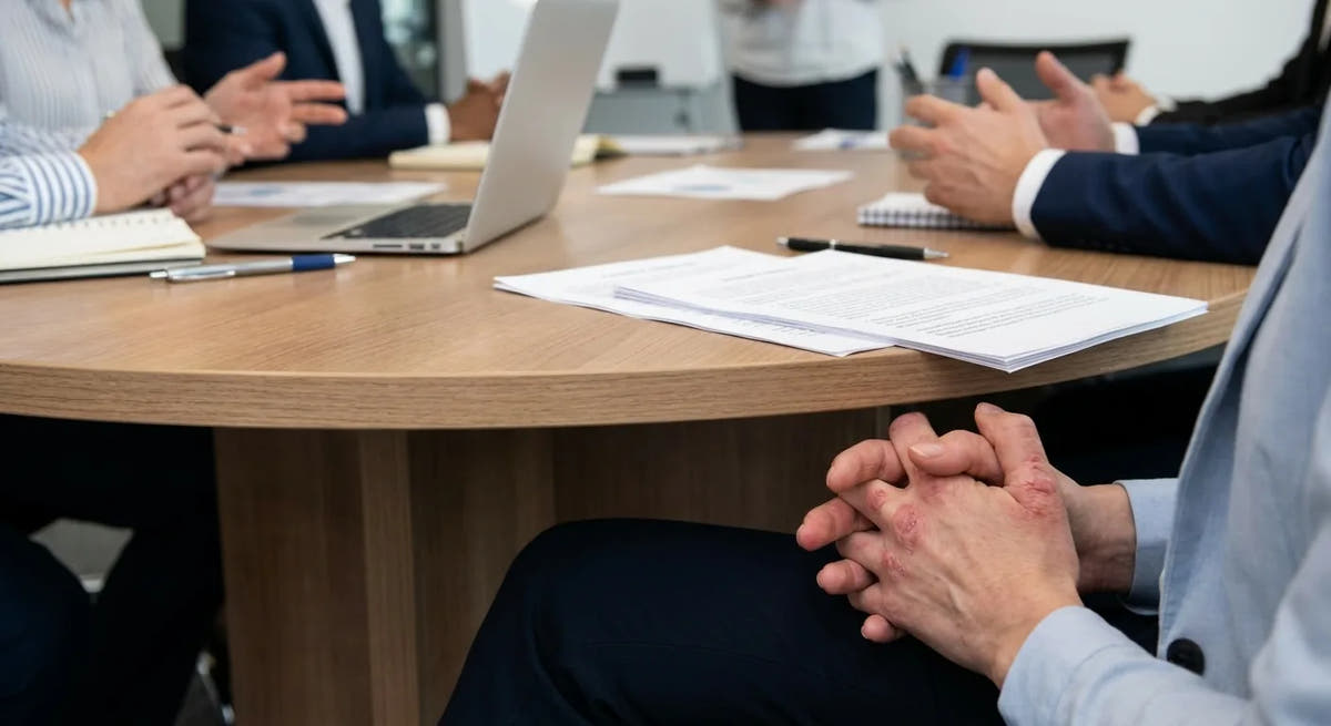 Les mains de Valérie posées sur la table pendant une réunion, doigts irrités, ordinateur et dossiers autour